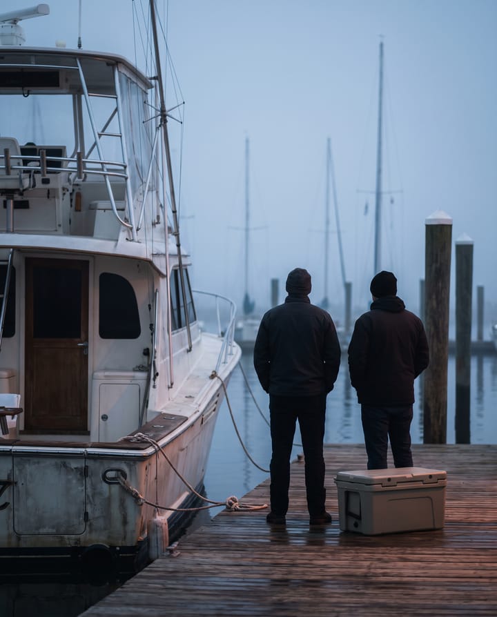 Three people in plain jackets and caps standing together on a misty marina dock at dawn, seen from behind, facing a sport-fishing boat tied alongside with a cooler and rods beside them.