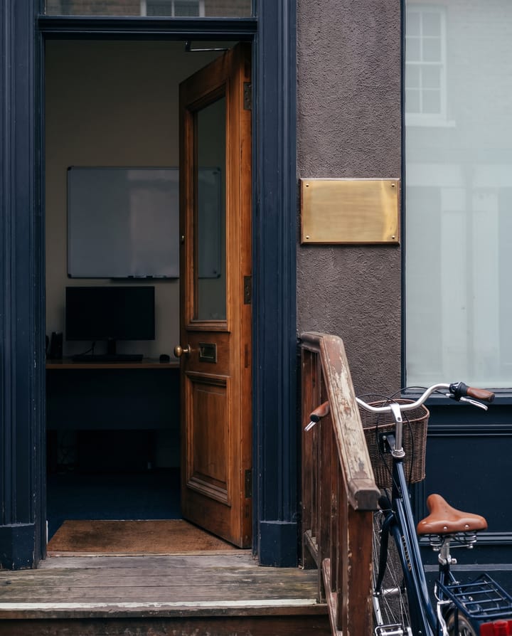 The open wooden door of a small office on a street, a blank brass plate on the wall beside it, a bicycle against the railing of the steps.