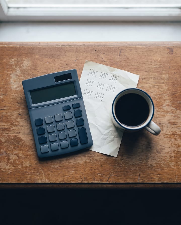 From above: a calculator with blank keys, a paper tally sheet of pencil marks in fives, and a mug of coffee on a wooden counter.