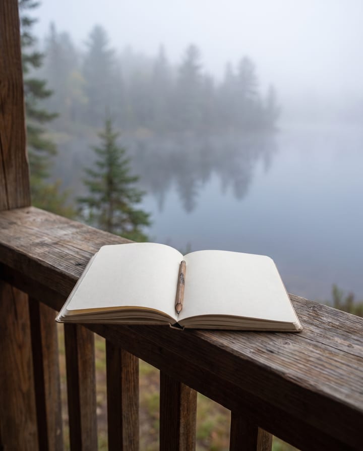 An open notebook with blank pages and a pencil in its gutter, on a porch rail above a misty lake and pines.