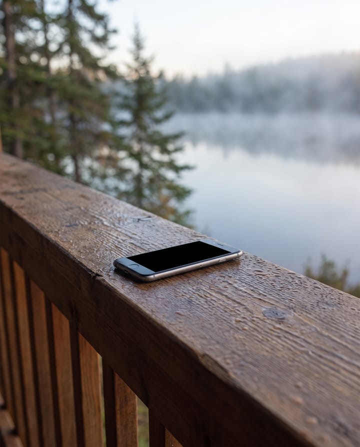 A phone with a dark screen lying on a wet porch rail, a misty lake and pines behind.