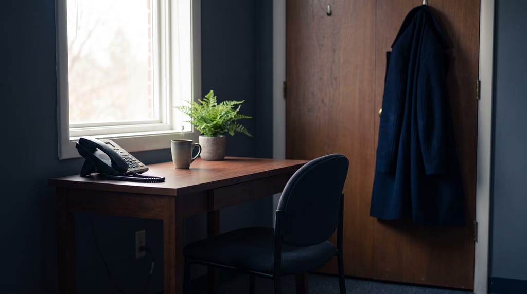 The corner of a small office: a plain desk against a window with a landline phone in its cradle, an empty chair pushed in, a mug and a potted fern, a coat on a hook by the door.