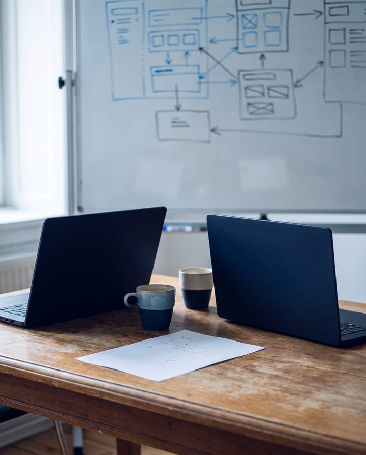 Two laptops and two coffee cups on one wooden table, a sheet of paper between them, a whiteboard of boxes and arrows behind.