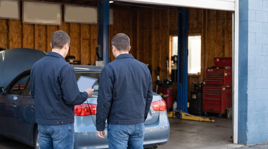 Two people seen from behind standing at the back of a car in the service lane of a repair shop, one holding a tablet, the open bay with a lift and tool chests beyond.