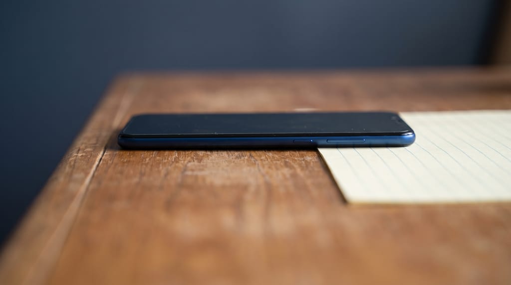 A phone with a dark screen lying on a wooden counter beside a sheet of ruled paper.