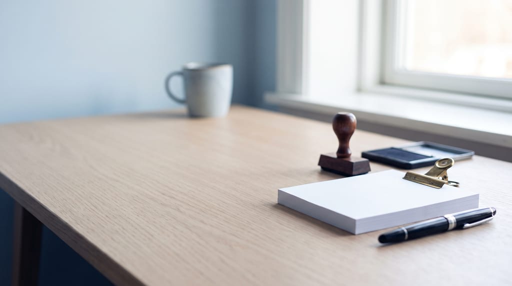 A pale oak desk by a window: a squared stack of blank paper held by a brass bulldog clip, a wooden rubber stamp and an ink pad, a black fountain pen, and a mug further back.