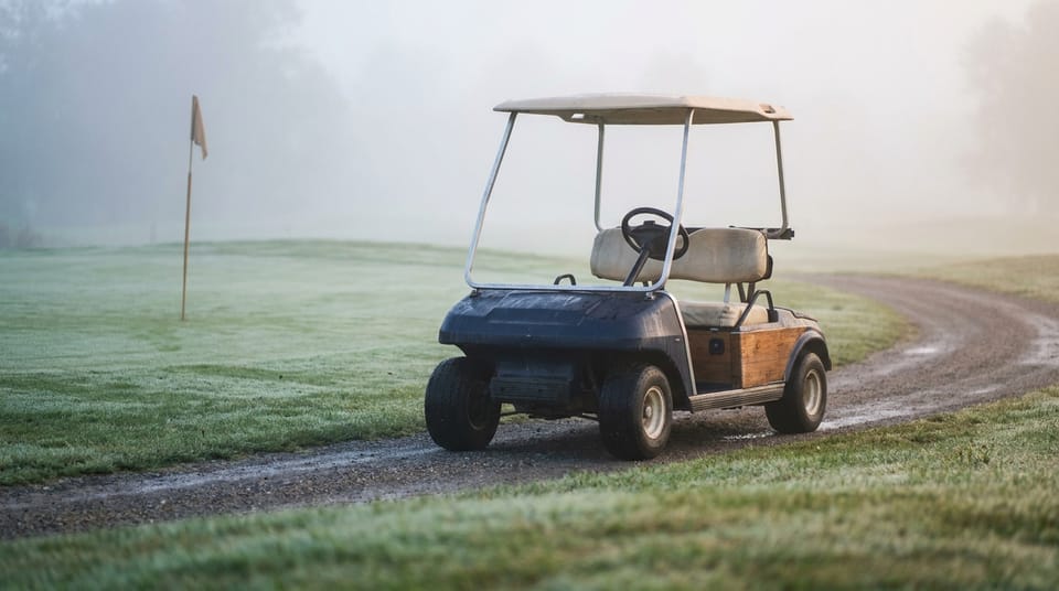 A golf cart on a curved cart path beside a green with a plain flagstick, morning mist.