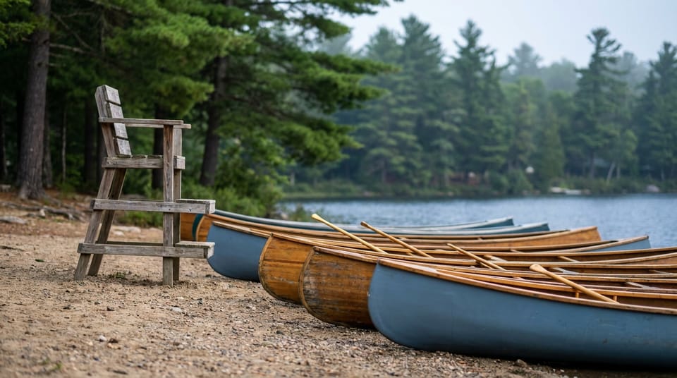 A row of canoes pulled up on a lake beach at a summer camp, an empty lifeguard chair, pines behind.