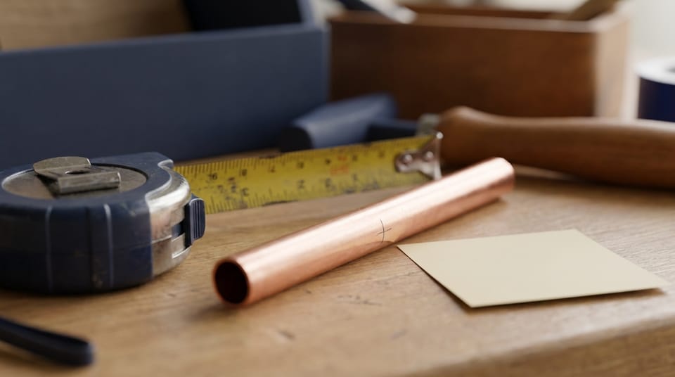 A tape measure extended along a fresh length of copper pipe on a workbench, a single pencil tick mark on the pipe, a blank sticky note beside it.