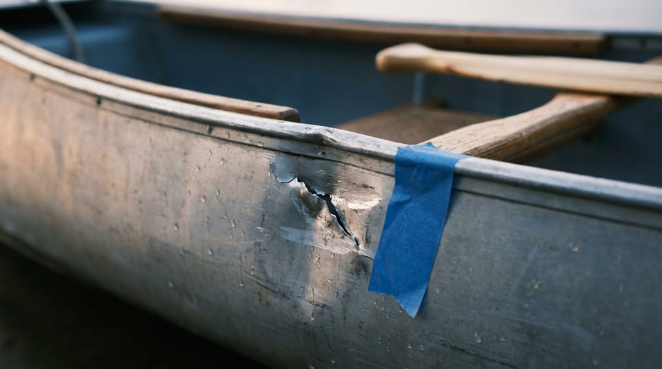 Close view of a fresh dent and scratch in the aluminium side of a canoe, a strip of blue painter's tape stuck beside it, water droplets on the metal.