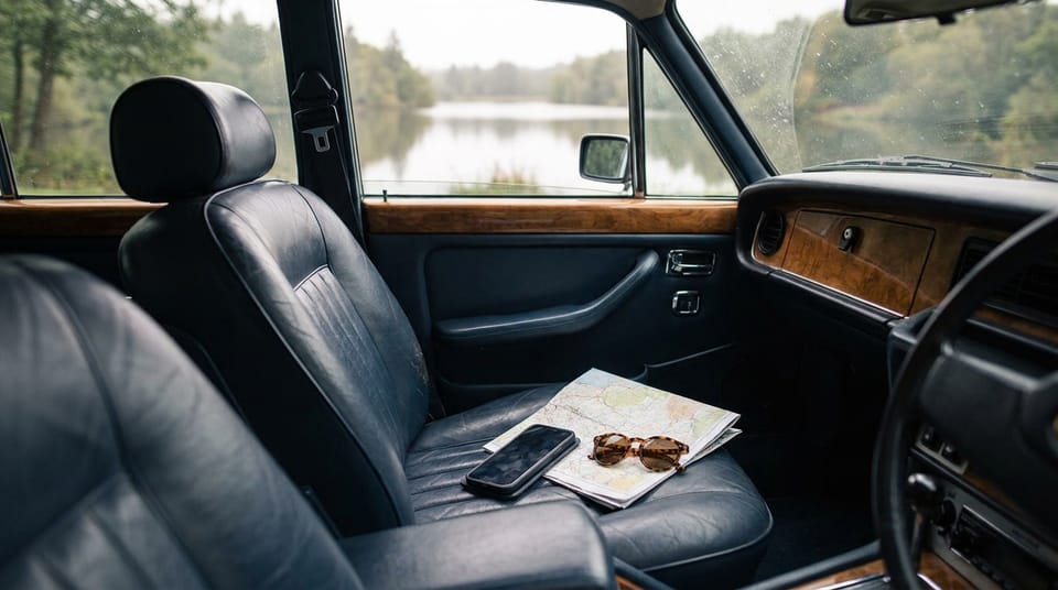 A phone lying on a folded paper map with a pair of sunglasses on the passenger seat of a car, a lake through the windows.