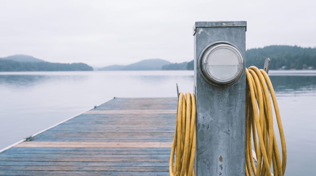 A marina power pedestal at the head of an empty slip with a coiled yellow shore-power cord, the finger pier running out to open water and low hills.