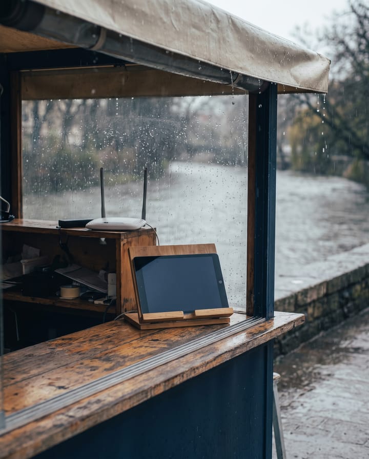 A tablet with a dark screen on a wooden stand under a riverside awning in the rain, a router on the shelf behind.