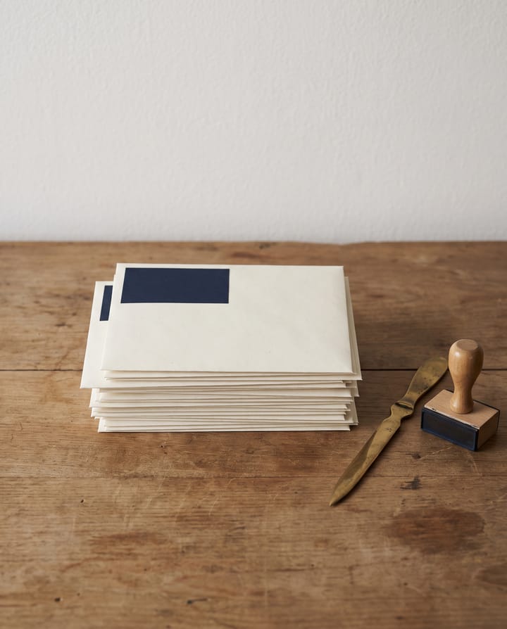 A stack of cream envelopes with blank navy labels on a wooden desk, a brass letter opener and a wooden rubber stamp beside them.