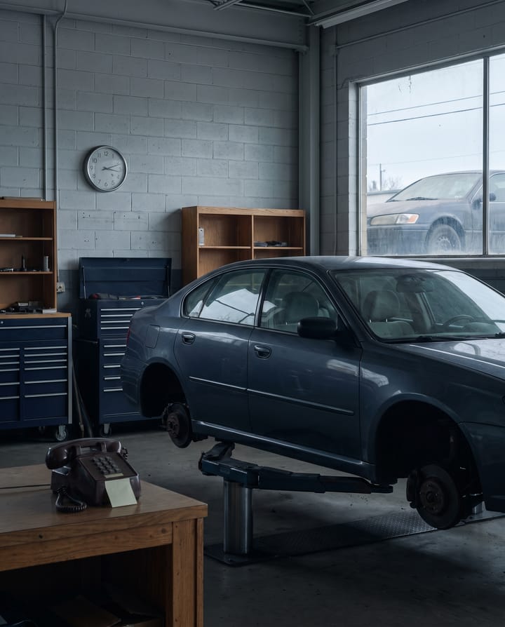 A plain grey sedan up on a lift with its front wheels off in a service bay, an old desk phone with a blank sticky note on the workbench, a wall clock, and a second dusty car through the window.