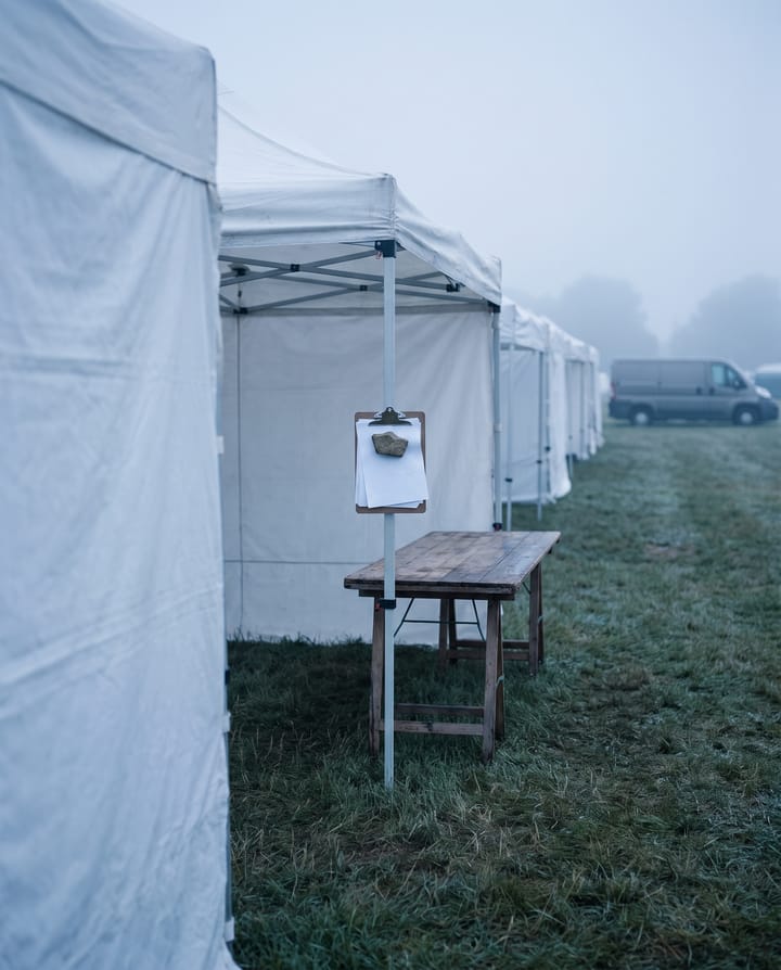 A misty festival field before the gates open: a row of white vendor tents, a clipboard of papers hanging from a tent pole over an empty folding table.