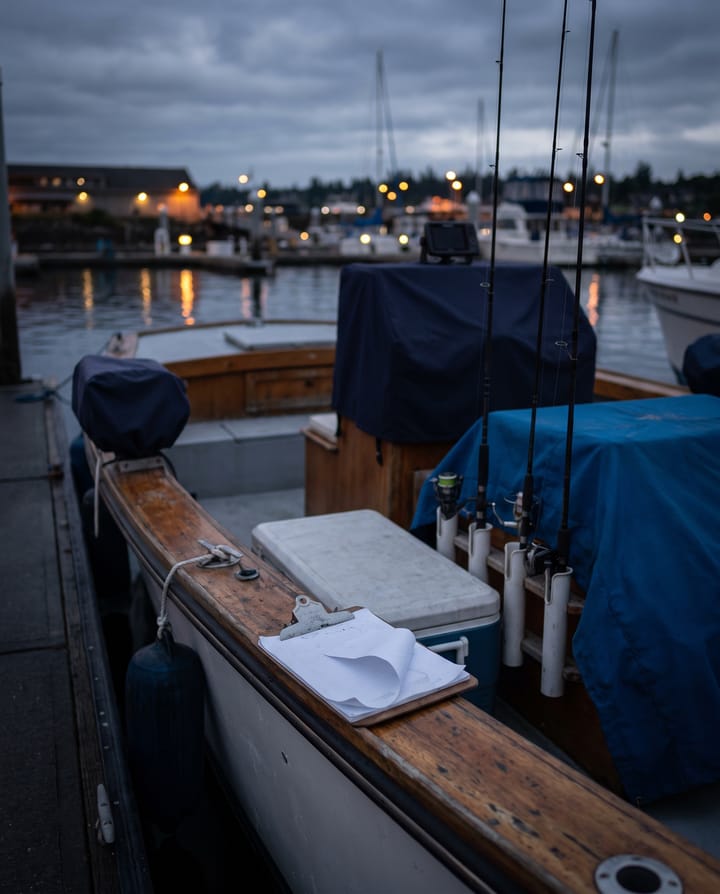 A charter boat's cockpit at the dock before dawn: rods in their holders, a cooler, a waterlogged clipboard of paper waivers curling on the gunwale, harbour lights beyond.