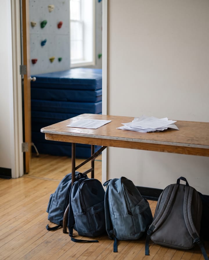 A folding table by a gym doorway with loose sheets of paper on it, four backpacks on the floor beneath, blue mats and a climbing wall behind.