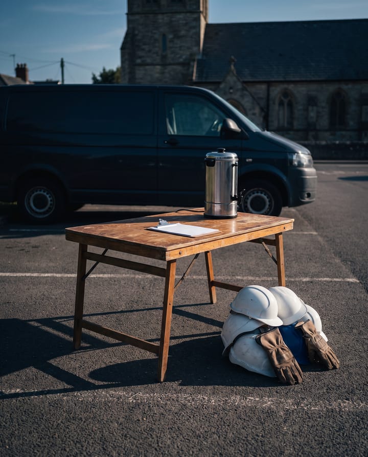 A folding table with a coffee urn and a clipboard in a church car park, hard hats and work gloves on the tarmac beneath, a dark van parked behind.