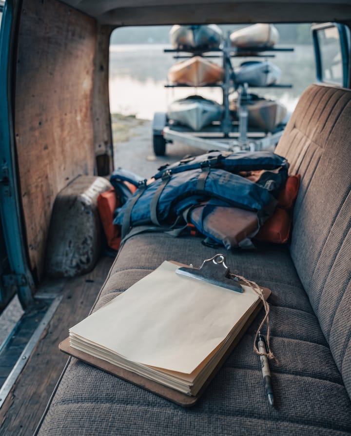 A clipboard of paper waivers with a pen on a string lying on the bench seat of an open van, life jackets piled behind and a trailer of kayaks by the water.