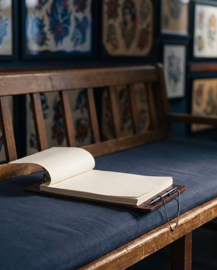 A clipboard holding a multi-page paper form lying open on the padded waiting bench of a tattoo studio, a pen on a bead chain, framed flash art on the wall behind.