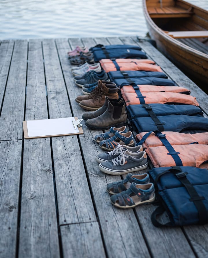 Shoes of different sizes, adults' and children's, lined up on a dock beside a row of life jackets, a clipboard on the planks.