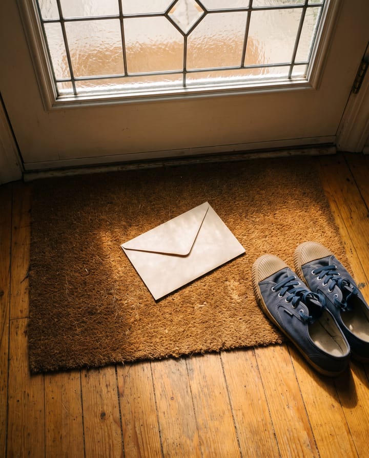 An unopened printed envelope lying on a doormat inside a front door, morning light through the glass, a pair of shoes beside it.